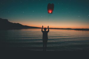 Person releasing a glowing lantern into the twilight sky over a calm lake, with stars and mountains in the background.
